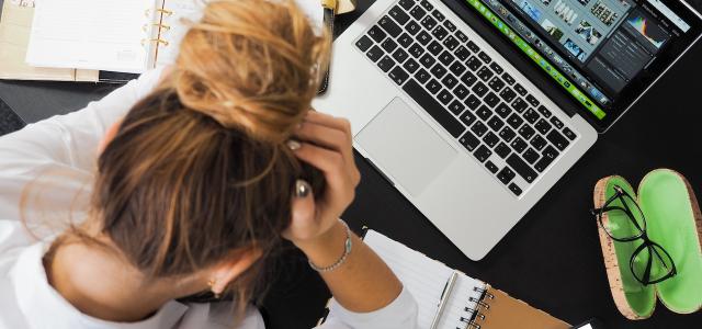 Top-down image of a woman sitting at a cluttered desk. She's holding her head and looking down.